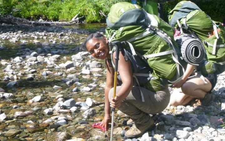 two teens wearing backpacks kneel beside a rocky stream on a outward bound course